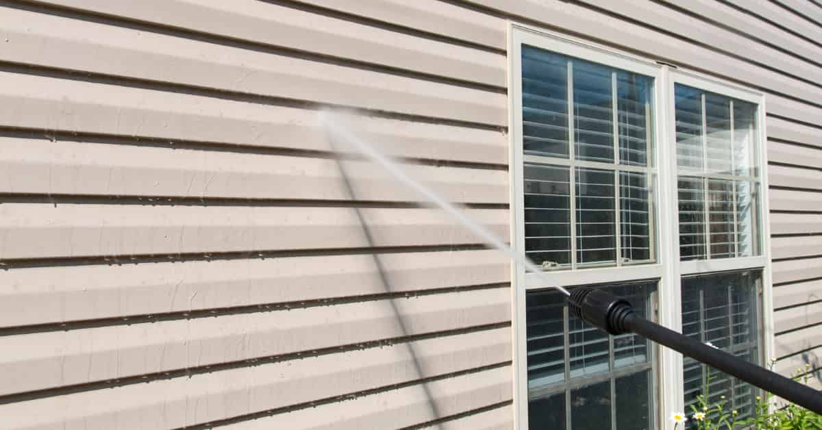 A high-pressure water cleaner is being used to wash the siding of a house, with water spraying across the surface.